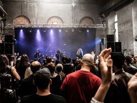 Metalband auf einer beleuchteten Bühne vor dicht gedrängtem Publikum in einer alten Industriehalle.