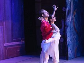 Tänzer in rotem Kostüm und Tänzerin in weißem Ballettkleid stehen gemeinsam in einer ausdrucksstarken Tanzpose auf einer blau-violett beleuchteten Bühne.