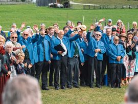 Männerchor in blauen Trachten singt vor Publikum auf einer Wiese.