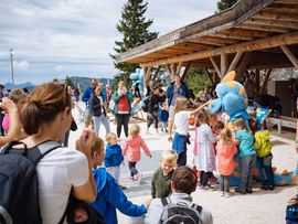 Viele Familien und Kinder versammeln sich bei einer Veranstaltung im Freien rund um eine blaue Dinosaurierfigur vor einer Berghütte.
