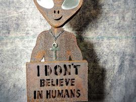Rostfarbene Metallskulptur einer außerirdischen Figur mit großen hellen Augen, Kette und einem Schild mit der Aufschrift „I Don't Believe in Humans“.