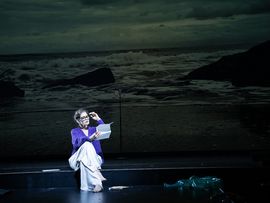 Schauspielerin in violetter Kleidung sitzt mit einem Blatt Papier vor einer Meeresprojektion.