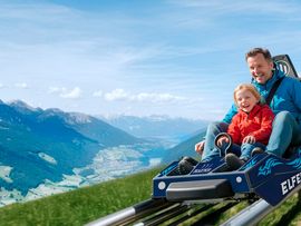 © Werbeagentur 38.5 Ein Vater und ein Kind fahren lachend mit dem Elfercoaster den Berg hinunter. Im Hintergrund erstreckt sich ein beeindruckendes Alpenpanorama mit Tal und schneebedeckten Gipfeln.