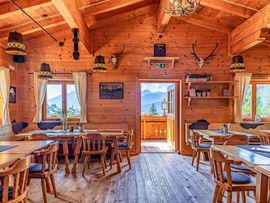 Gemütlicher Gastraum der Enzianhütte mit Wänden, Decke und Fußboden aus Holz. Holztische und Stühle stehen unter rustikalen Lampen; durch die Fenster und die geöffnete Tür bietet sich ein Blick auf die umliegende Berglandschaft.
