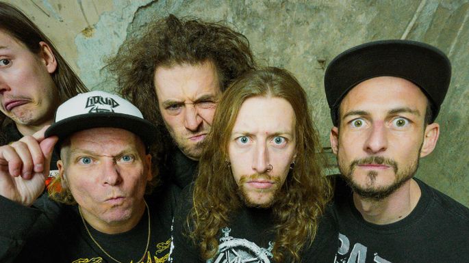 Fünf Musiker posieren gemeinsam für ein Gruppenfoto und blicken mit auffälligen, teils humorvollen Gesichtsausdrücken in die Kamera. Die Band trägt überwiegend schwarze Kleidung und steht vor einer rustikalen, urban wirkenden Wand.