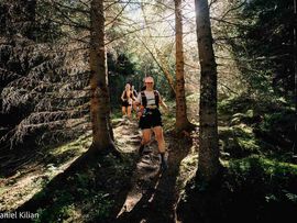 Zwei Trailrunnerinnen laufen auf einem schmalen Waldweg durch einen sonnendurchfluteten Nadelwald.