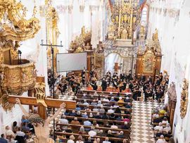 Blick von der Empore auf das Festkonzert in der barocken Stiftskirche Stams. Das Orchester spielt vor zahlreichen Besucherinnen und Besuchern in der voll besetzten Kirche.