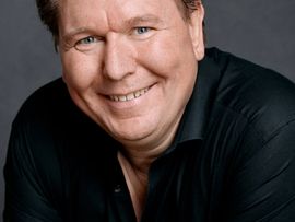 ©ChristianPalm Porträt des Tenors Thomas Heyer. Er lächelt direkt in die Kamera und trägt ein schwarzes Hemd vor grauem Hintergrund.