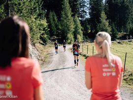 Mehrere Trailrunner laufen auf einem breiten Schotterweg durch eine sonnige Waldlandschaft, beobachtet von Zuschauenden im Vordergrund.