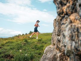 Eine Trailrunnerin läuft über einen grasbewachsenen Berggrat entlang einer markanten Felswand.