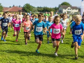 Eine Gruppe von Kindern startet gemeinsam zu einem Lauf über eine Wiese. Im Vordergrund laufen mehrere Kinder mit Startnummern, im Hintergrund folgen weitere Teilnehmende.