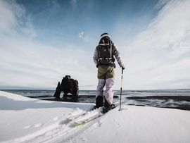 Eine Person auf Tourenski blickt über eine verschneite Küstenlandschaft mit schwarzem Felsbogen, Meer und weitem Himmel.