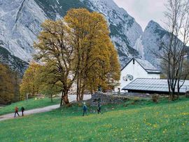 Der Alpengasthof St. Magdalena liegt auf einer grünen Wiese am Fuß einer imposanten, teils schneebedeckten Berglandschaft. Große Laubbäume stehen vor dem weißen Gebäude, mehrere Wanderer sind auf dem Weg unterwegs. Es ist Herbst.