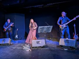 Die Musikgruppe Schwarzes Edelweiss bei einem Live-Auftritt auf einer Bühne. Eine Sängerin steht in der Mitte, begleitet von einem Gitarristen und einem Bassisten.