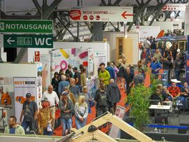 Besucherinnen und Besucher flanieren durch die Ausstellungsbereiche einer stark frequentierten Messehalle.
