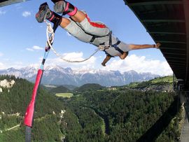 „Bungy-Springer stürzt kopfüber von einer hohen Brücke und hängt am elastischen Seil über einem bewaldeten Alpental.“