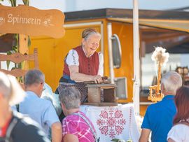 Eine ältere Frau in traditioneller Tracht zeigt an einem Marktstand das Spinnen von Wolle. Mehrere Besucherinnen und Besucher beobachten die Handarbeit.