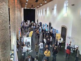 Blick von oben auf eine Veranstaltungshalle mit zahlreichen Ausstellerständen und Besucher:innen.