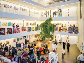 Mehrstöckiger Ausstellungsraum im Atrium Amras mit farbenfrohen Gemälden, Besuchern und einem großen Baum in der Mitte der Halle.