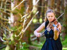 Eine Geigerin in einem langen schwarzen Kleid spielt mit geschlossenen Augen Violine auf einer moosbedeckten Waldlichtung. Zwischen den hohen Bäumen fällt Sonnenlicht durch die Äste und schafft eine ruhige, stimmungsvolle Atmosphäre.