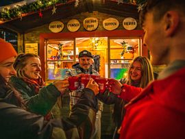 Gruppe stößt mit Heißgetränken an einem Stand beim Imster Weihnachtsmarkt freudig miteinander an.