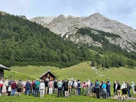 „Menschen versammeln sich vor einer kleinen Almkappelle zur Bergmesse auf der Eng Alm, umgeben von einer beeindruckenden alpinen Berglandschaft.“