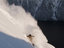 Eine Person fährt durch tiefen Pulverschnee einen steilen Berghang hinab, während im Hintergrund dunkles Wasser und verschneite Berge liegen.