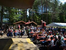 Besucherinnen und Besucher feiern im Festgelände, während Trachtler auf Bänken tanzen und Kindergruppen traditionelle Darbietungen zeigen.