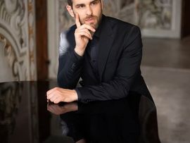 Porträt des Dirigenten Elias Praxmarer in elegantem schwarzen Anzug, sitzend an einem Flügel in einem barocken Saal. Sein Spiegelbild ist auf der glänzenden Oberfläche des Klaviers zu sehen.