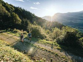 Zwei Trailrunner laufen bei tiefstehender Sonne auf einem Bergweg mit Blick über ein bewaldetes Tal.