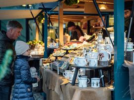 Kind und erwachsene Person betrachten Tassen, Schmuck und Geschenkartikel an einem weihnachtlich dekorierten Marktstand.