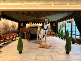 Ein stilvoll gestalteter Innenraum mit einer historischen Pferdekutsche und einem weißen Pferd als Ausstellungsstück. Die elegante Einrichtung mit Sitzgelegenheiten, dekorativen Pflanzen und Wandgemälden vermittelt ein hochwertiges Museumserlebnis.