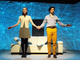 Mann und Frau vor abstraktem Bühnenhintergrund blicken händehaltend Richtung Publikum