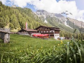 Die Schlickeralm liegt auf einer grünen Almwiese vor einer bewaldeten Berglandschaft. Das Gebäude verfügt über eine große Sonnenterrasse mit roten Sonnenschirmen; im Hintergrund ragen markante Felsgipfel auf.