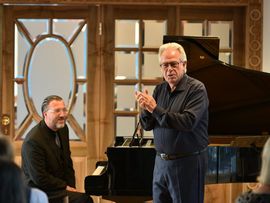 Ein Gesangsdozent erläutert vor Publikum musikalische Inhalte und gestikuliert dabei mit den Händen. Neben ihm sitzt ein Pianist am geöffneten Konzertflügel und verfolgt aufmerksam die Ausführungen.