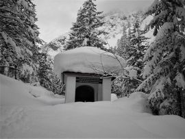 Eine kleine Kapelle liegt tief verschneit in einem winterlichen Bergwald. Auf dem Gebäude und den umliegenden Tannen liegt eine dicke Schneedecke, im Hintergrund erhebt sich ein verschneiter Berghang.