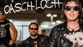 Bandfoto bei Nacht: mehrere Männer in Lederjacken und Sonnenbrillen, vorne ein Mann mit kleinem weißen Hund im Arm; darüber der Schriftzug „Oaschloch“.