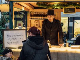 Person bedient einen festlich geschmückten Marktstand, während Besucherinnen und Besucher über einen stimmungsvollen Weihnachtsmarkt schlendern.