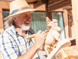 Ein älterer Mann mit Hut und kariertem Hemd schnitzt mit einem Werkzeug eine Holzfigur an einem Marktstand.
