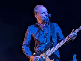 Ein Bassist der Gruppe Schwarzes Edelweiss spielt auf einer Bühne unter blauem Bühnenlicht und steht dabei am Mikrofon.