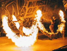 Zwei Künstlerinnen präsentieren eine Feuershow auf der Bühne. Mit brennenden Feuergeräten erzeugen sie große Flammen vor der Kulisse von Masters of Dirt.
