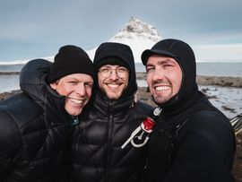 Drei warm eingepackte Personen stehen an einer verschneiten Küste vor einem markanten, schneebedeckten Berg.