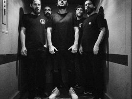 Schwarzweißfoto von fünf Männern, die gemeinsam in einem engen Aufzug stehen. Alle tragen dunkle Kleidung; die Aufnahme wirkt wie ein Bandfoto vor einem Konzert.