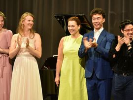 Mehrere Gesangsstudierende und Lehrende stehen nach dem Konzert gemeinsam auf der Bühne vor einem Flügel und applaudieren lächelnd dem Publikum.