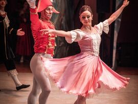 Ballettszene aus dem Nussknacker mit einer Tänzerin in einem rosafarbenen Kleid und einem Tänzer im Kostüm des Nussknackers auf einer beleuchteten Bühne.