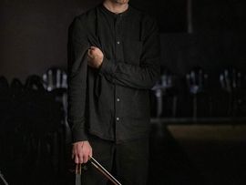Portrait eines Geigers mit zusammengebundenen Haaren und schwarzer Kleidung. Er steht in einem dunklen Innenraum und hält eine Violine und einen Geigenbogen in der Hand. Im Hintergrund sind unscharf Stühle eines Konzertsaals zu erkennen.