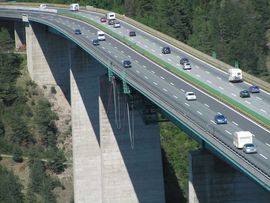„Mehrspurige Autobahn mit Autos und Lastwagen auf einer hohen Brücke über einem bewaldeten Alpental.“