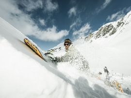 Eine Person fährt auf Skiern durch tiefen Pulverschnee in einer steilen, von schneebedeckten Felsen umgebenen Berglandschaft.