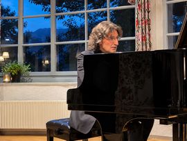 Ein Pianist mit schulterlangem, grauem Haar spielt an einem schwarzen Konzertflügel in einem Raum mit großen Fenstern und gemusterten Vorhängen.
