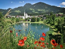 „Blick über einen See auf den Ort Reith im Alpbachtal mit Kirche, grünen Wiesen und bewaldeten Bergen im Hintergrund.“